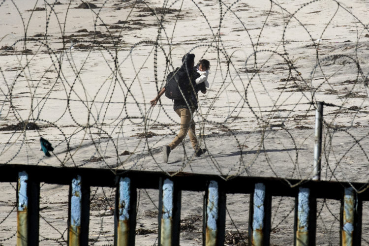 TIJUANA, MEXICO  03-13-2019: A 22-year old Honduran man carries his young daughter in his arms as he runs down the beach on the U.S. side of the border wall, after he squeezed through pillars and fencing at La Playa in Tijuana. The man dramatically sprinted down the beach towards San Diego. Remarkably, he was not captured, and is currently living with family elsewhere in the U.S.