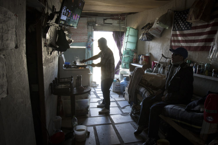 TIJUANA, MEXICO  03-12-2019: Two migrants cook food  on open stove and watch TV in the midday light in a small boarding room in La Playa, near the beach and border wall in Tijuana.