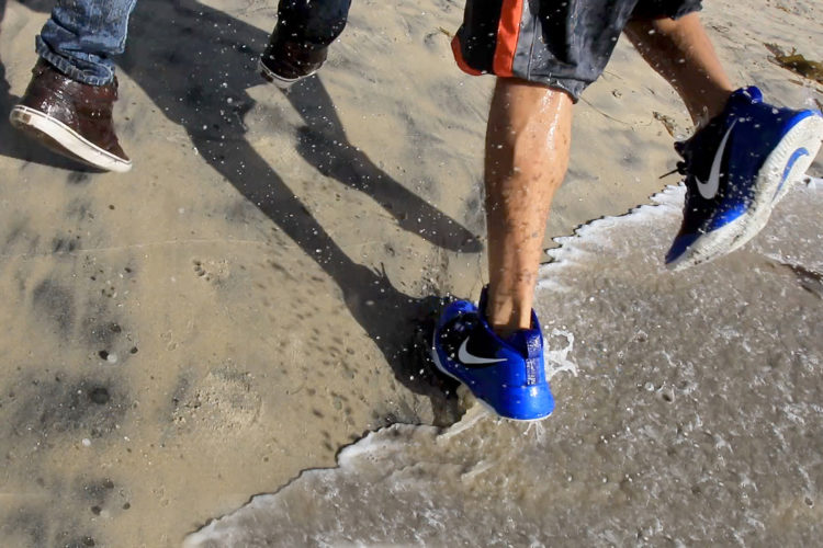 TIJUANA, MEXICO  03-14-2019: Two migrants run in the surf after they squeezed through an opening in the border fence on the beach running past a CBP agent and ran north along the beach into the United States. They were later captured by United CBP agents, who said they arrested about 52 migrants, including 23 men, between the ages of 18 to 53, 12 women aged 21 to 50, and 17 minors between the ages of 1 and 14 years old. They were mostly from Honduras and came to Tijuana in a caravan. As they broke through the fence and ran down the beach, some dropped clothes and shoes from the backpacks, as some people on the Mexico side cheered them on.