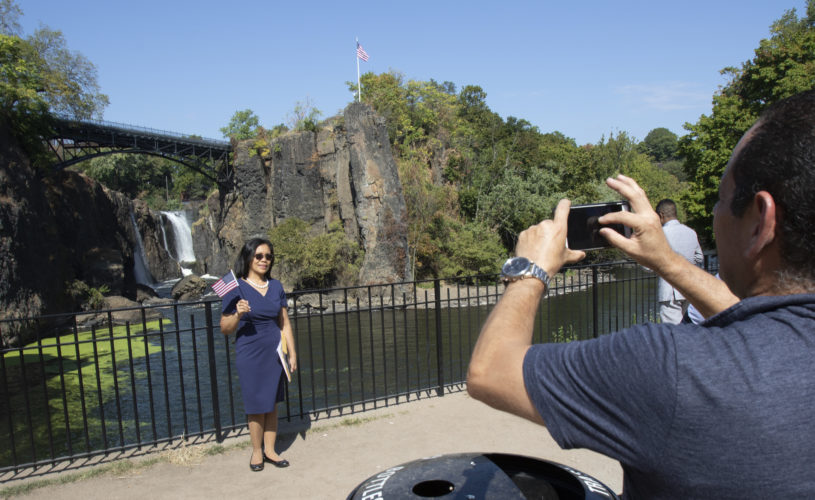 PATERSON, NJ  10-02-2019 NATURALIZATION CEREMONY AT GREAT FALLS:  Marlin Geron Rivas waves the U.S, flag while having her photo taken by the Great Falls, after taking the Oath of Allegiance to the United States. She is from the Philippines. The Paterson Great Falls National Historical Park (NPS), in partnership with U.S. Citizenship and Immigration Services (USCIS), held a naturalization ceremony in the park’s new amphitheater where 40 new citizens, mostly residents of Paterson, took the Oath of Allegiance to the United States.